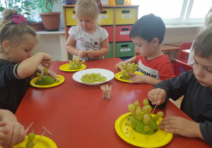 dzieci nakłuwają winogrona na wykałaczki wkłute w połowę gruszki umieszczonej na żółtych talerzykach