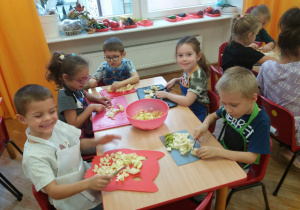 dzieci w fartuszkach kroją na deskach owoce i wrzucają do miski na środku stolika