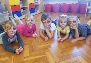 dzieci z pomalowanymi twarzami na wzór mordki pieska pozują do zdjęcia leżąc na brzuchu na podłodze w sali przedszkolnej