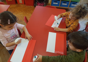 dzieci przy stoliku sklejają paski białego i czerwonego papieru i robią flagę Polski