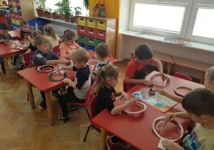 dzieci przy stolikach malują farbą papierowe obręcze z przyklejonymi uszami