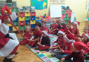 dzieci z grupy VIII grają Mikołajowi na drewienkach, siedząc na dywanie przed Mikołajem