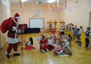 dzieci siedzą z prezentami i żegnają Mikołaja z zarzuconym na plecy workiem i trzymającym laskę