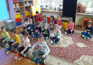 dzieci siedzą na dywanie i grają na bębenkach z puszek pałeczkami z korków i patyczków do szaszłyków