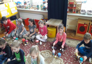 dzieci siedzą obok żółtej tablicy i grają na bębenkach z puszek pałeczkami z korków i patyczków do szaszłyków