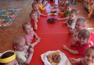 dzieci przy długim czerwonym stole siedzą i częstują się ciasteczkami