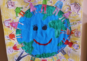 plakat grupy V - Ziemia pomalowana farbami a na niej stoją dzieci narysowane kredkami i naklejone na Ziemię z wypisanymi imionami