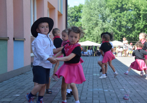 "Polka cowboyka" taniec dzieci z grupy VIII w parach - dziewczynki w różowych spódniczkach i chustkach oraz w czarnych podkoszulkach a chłopcy w krótkicj spodenkach, biłych koszulach i kapeluszach tańczą w parach po kole