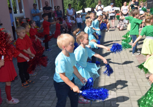 dzieci z grupy IV - tańczą z pomponami, ubrani w trzech kolorach: zielonym, czerwonym i niebieskim ustawieni w rzędach