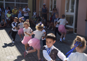 dzieci z grupy VI poskakują w parach, dziewczynki ubrane w białe bluzki i różowe, tiulowe spódniczki a chłopcy w białych koszulach , ciemnych spodniach i srebrzysto - szarych frakach