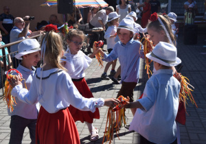 dzieci z grupy III tańczą w kołach - dziewczynki są w białych bluzkach i hiszpańskich spódnicach oraz z czerwonym kwiatem we włosach, natomiast chłopcy w białych kapeluszach i koszulach oraz ciemnych spodniach trzymają się za ręce i podtrzymują małe tamburyna ozdobione kolorowymi wstążkami