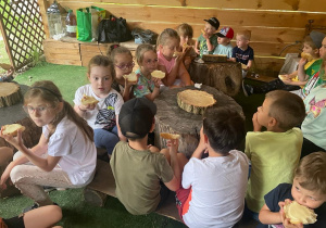 dzieci siedzą przy stolikach zpni drzewa i jedzą bułki z serem