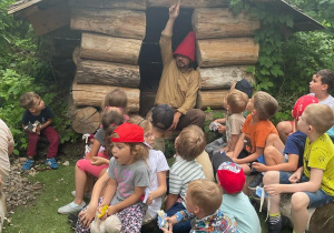 dzieci siedzą przed domkiem z bali drzewa i słuchają opowiadania skrzata w czerwonej czapce