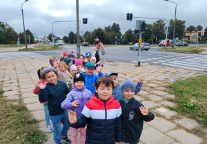 przedszkolaki stoją na chodniku przed skrzyżowaniem ulic i machają