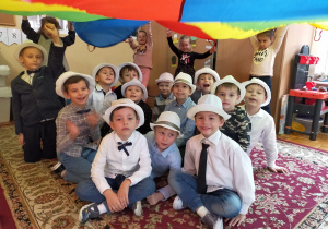 chłopcy z grupy III w białych kapeluszach siedzą na dywanie pod kolorową chustą, którą trzymają dziewczynki
