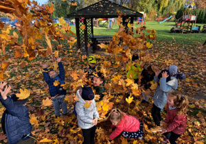 dzieci w ogrodzie przedszkolnym bawią się kolorowymi liściami podrzucając je do góry