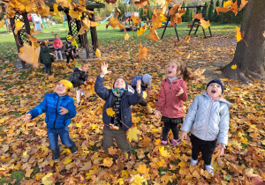 dzieci w ogrodzie przedszkolnym bawią się kolorowymi liściami podrzucając je do góry