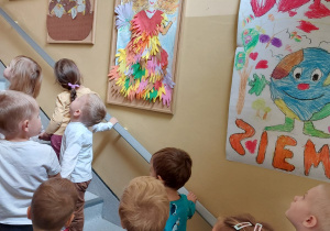 maluszki idą po schodach przedszkolnych i oglądają galerie prac wywieszonych na przedszkolnym korytarzu - planetę Ziemię, portret pani Jesieni, kosz z grzybami, kosz z owocami