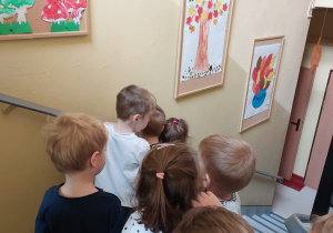 maluszki idą po schodach przedszkolnych i oglądają galerie prac wywieszonych na przedszkolnym korytarzu - grzyby jadalne i niejadalne, jesienne drzewo, wazon z kolorowymi liśćmi