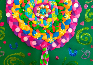 praca plastyczna wykonana z kolorowej plasteliny, posplatanej w warkocze, które ułożone w okręgi i ozdobione cekinami w kształcie kwiatów i kropek tworzą lizak na nużce z kolorowego drucika