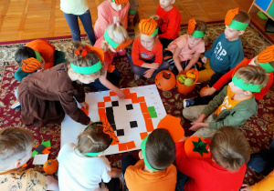 przedszkolaki układają na dywanie obrazek dyni z kolorowych kwadratów wg kodu liczbowo - literowego
