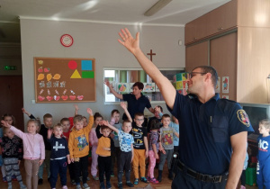 funkcjonariusz Straży Miejskiej stoi przed dziećmi i demonstruje im prawą stronę podnosząc prawą rękę do góry a dzieci powtarzają jego ruch