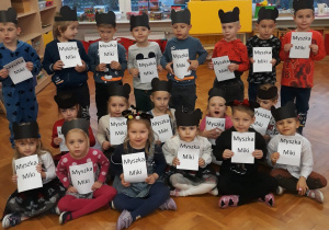 dzieci z grupy VI pozują do zdjęcia grupowego w czarnych opaskach z uszami Myszki Miki a w dłoniach trzymają kartkę z wydrukowanym napisem Myszka Miki