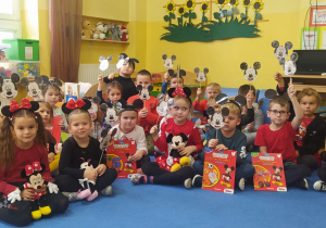 dzieci z grupy VIII pozują do zdjęcia grupowego w czarnych opaskach z uszami Myszki Miki i książeczkami, maskotkami Miszki Miki oraz głową Myszki Miki zamocowaną na patyczku