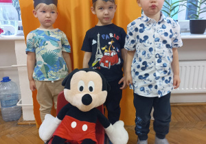 trzech chłopców stoi wokół dużej maskotki Myszki Miki siedzącej na krzesełku