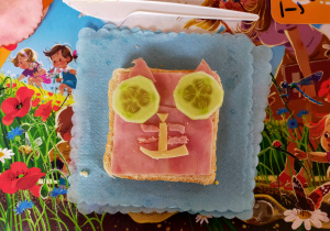 kanapka wykonana przez dziecko na niebieskiej serwetce - kotek - na kwadratowym chlebie wędlina, oczy z plastrów ogórka, nos i usta z cienkich kawałków zółtego sera, wąsiki i uszy z kawałków wyciętych z wędliny