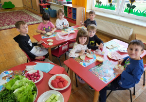 przedszkolaki siedzą przy stolikach i smarują kanapki masłem a obok na stoliku warzywa, ser, wędlina pokrojone i ułożone na talerzykach do komponowania zdrowych kanapek