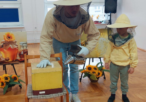 pszczelarz i chłopiec w kapeluszach z siatką stoją obok skrzynki a pszczelarz demonstruje w jaki sposób okadza pszczoły w ulu