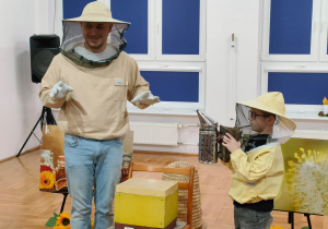chłopiec w pszczelarskim kapeluszu trzyma dymiarkę pszczelarską stojąc obok pszczelarza i ula