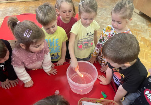 dzieci stoją wokół pojemnika z wodą a dziewczynka trzyma zapaloną świeczkę i leje wosk na wodę