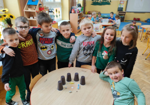 przedszkolaki stoją wokół okrągłego stolika, na którym stoją kubki oraz przedmioty, które będą schowane pod nimi