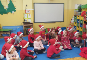 przedszkolaki w czerwonych czapkach siedzą przed Mikołajem i słuchają jego opowiadania