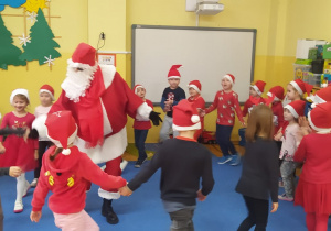 przedszkolaki tańczą w kole trzymając się za dłonie a w środku tańczy Mikołaj