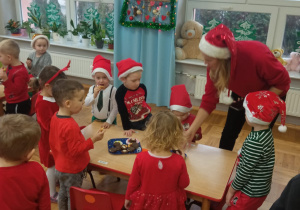 nauczycielka w czerwonej czapce zaprasza dzieci do stolików na słodki poczęstunek