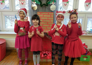 dziewczynki w czerwonych ubraniach stoją przed kominkiem i pokazują drobne, słodkie upominki od Mikołąja