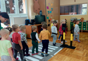 przedszkolaki przechodzą parami po pasach naklejonych na podłodze w sali i stojącymi sygnalizatorami świetlnymi