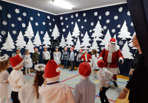 dzieci tańczą z Mikołajem w kole na tle świątecznej dekoracji - białych choinek i śniegowych gwiazdek