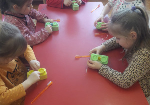 dzieci siedząc przy stoliku uczą się czyścić szczeliny międzyzębowe, wykorzystują kolorowe klocki zaklejone plasteliną i sznurek
