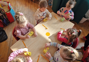 dziewczynki siedząc przy stoliku uczą się czyścić szczeliny międzyzębowe, wykorzystują kolorowe klocki zaklejone plasteliną i sznurek