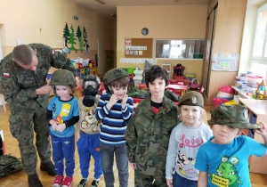 żołnierz pomaga założyć chłopcu hełm wojskowy a obok stoja dzieci ubrane w maskę, beret, czapkę i mundur