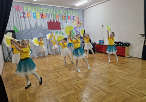 dziewczynki w żółtych koszulkach i biało zielonych spódniczkach tańczą z kwiatami podnosząc je w górę