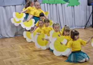 dziewczynki w żółtych koszulkach i biało zielonych spódniczkach tańczą z kwiatami stojąc jedna za drugą w rzędzie