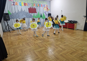dziewczynki w żółtych koszulkach i biało zielonych spódniczkach tańczą z kwiatami idąc po kole