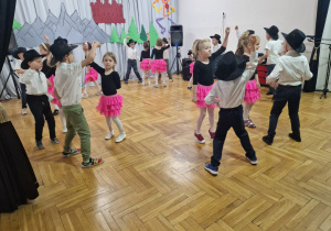 dziewczynki w różowych spódniczkach tańczą z chłopcami w czarnych kapeluszach stojąc w parach na obwodzie koła machając jedną ręką w górze