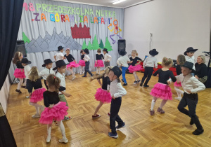 dziewczynki w różowych spódniczkach tańczą z chłopcami w czarnych kapeluszach podskakując w parach na obwodzie koła