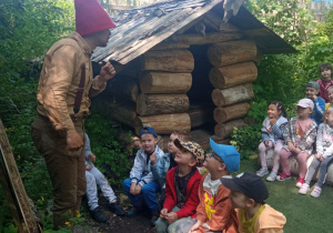 przedszkolaki słuchają opowiadania Skrzata siedząc przed drewnianym domkiem